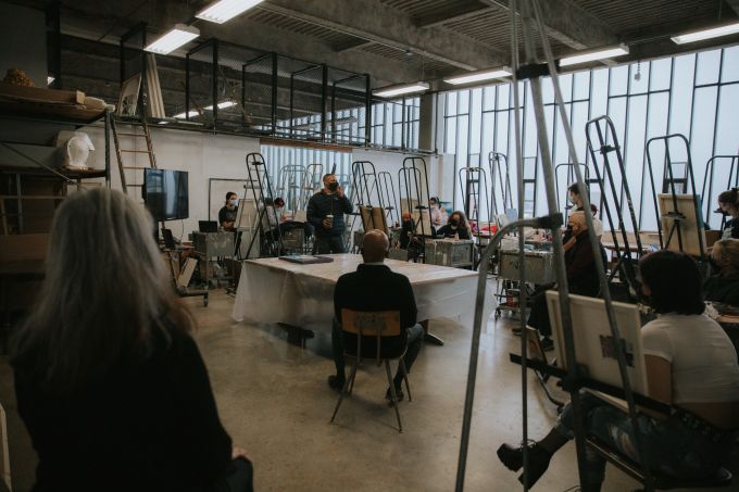 Around 15 students and professors, seated throughout a large art studio listening to a black man wearing a surgical mask and holding a coffee cup.