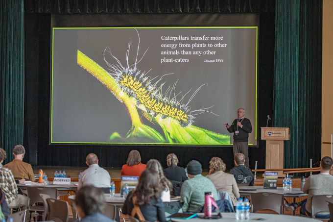 A man stands on a stage at right speaking to seated guests with an image of a caterpillar projected behind him