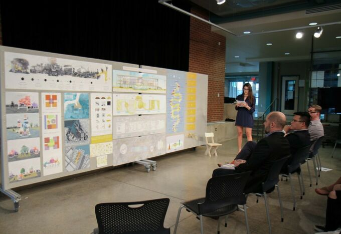 Female architecture student at far right refers to her presentation boards to her right with the jury in front of her.