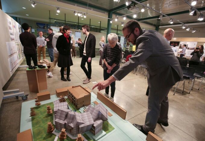 Male architecture student in the foreground points to his model on the floor as people watch him.