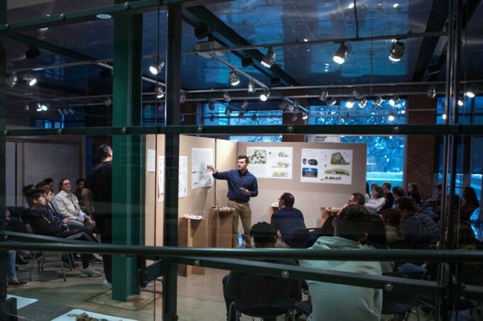 An architecture student explaining his project displayed on a wall board in front of faculty and staff during a critique.