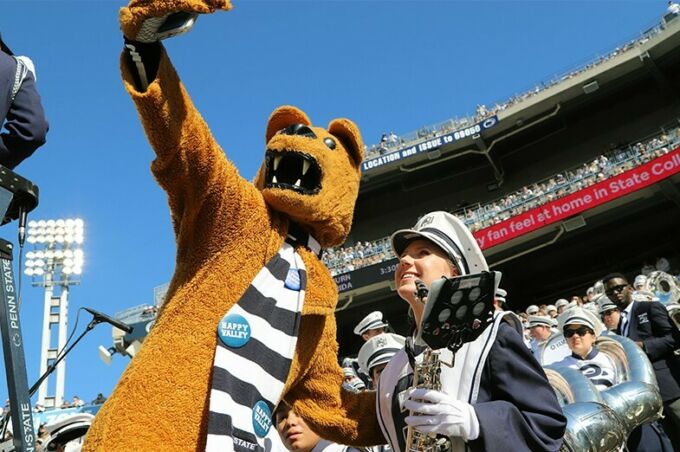 During college game day, a blue band member takes a selfie with the Nittany Lion during the football game.