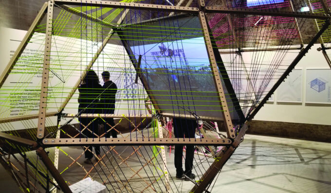 View through a faceted polygonal structure with tensioned support strings into an exhibition area with people in the background examining content posted on walls.