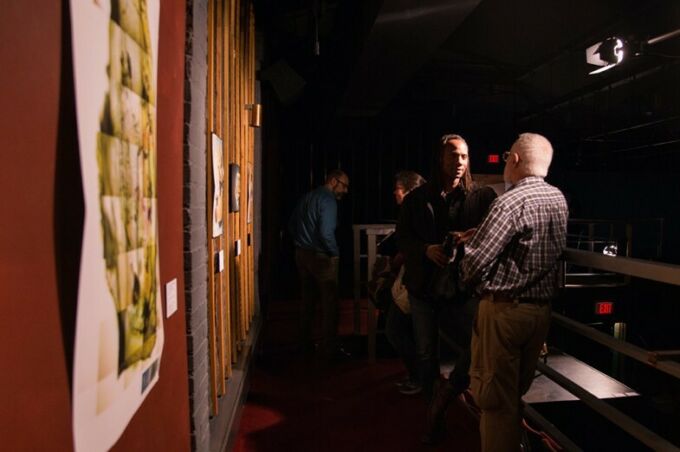 Visitors talking in shadows along a mezzanine-level walkway; close-up of art pieces hung on wall.