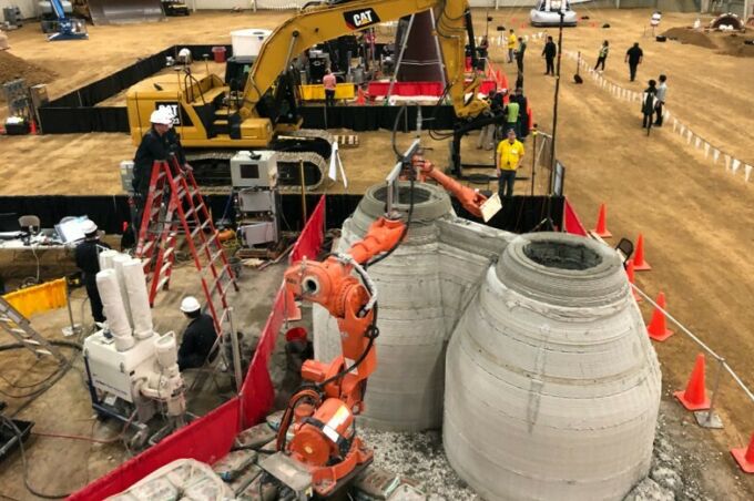 Inside a huge warehouse, large Caterpillar excavator equipment lifts industrial robots into place to make scale models of human-habitable enclosures from printed concrete.