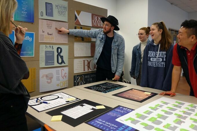 A graphic design student presents his typography work displayed on a wall during studio class.