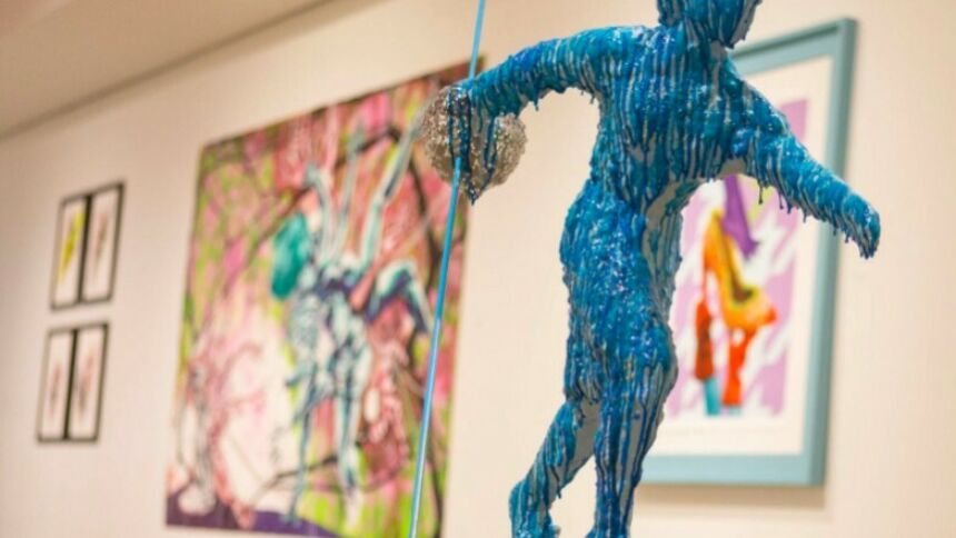 Blue-wax-drip-coverd figure on a pedestal in the foreground, with brightly colored, but focus-blurred paintings and framed artworks hung on a wall in the background.