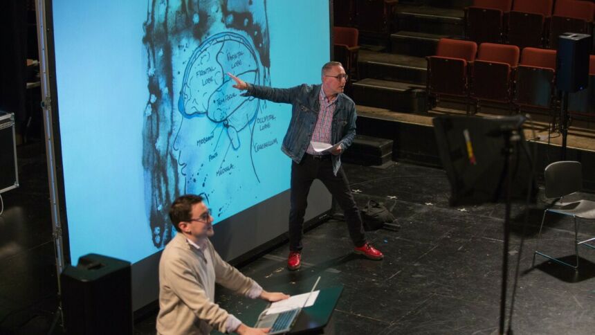 Bill Doan gesturing toward a projected illustrated diagram of a human brain. A man is standing in the foreground at a podium with a laptop.