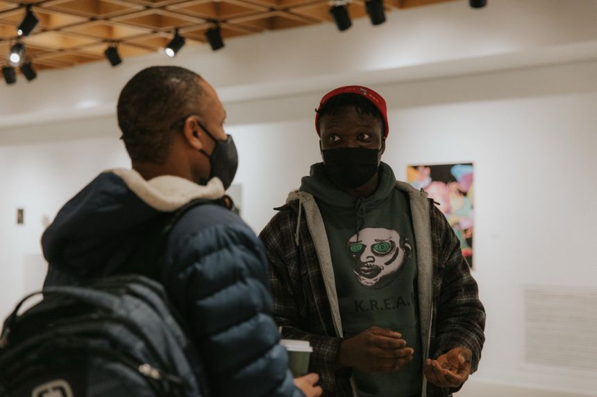 Closeup of a black student artist, wearing a sweatshirt with a caricature illustrated head on the chest, facing the camera and speaking to another black man wearing a blue, hooded puffy jacket.