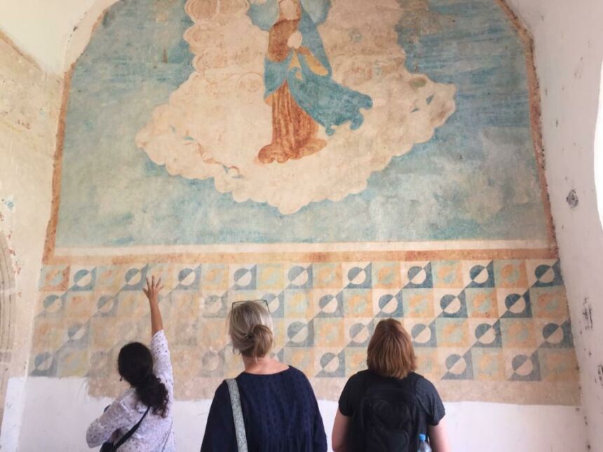 Three women stand facing a mural and one points at it.