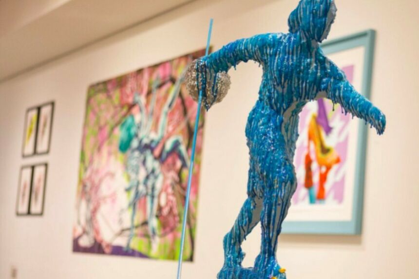 Blue-wax-drip-coverd figure on a pedestal in the foreground, with brightly colored, but focus-blurred paintings and framed artworks hung on a wall in the background.