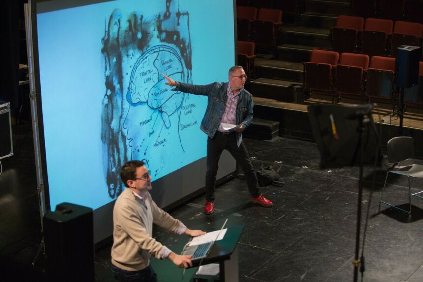 Bill Doan gesturing toward a projected illustrated diagram of a human brain. A man is standing in the foreground at a podium with a laptop.