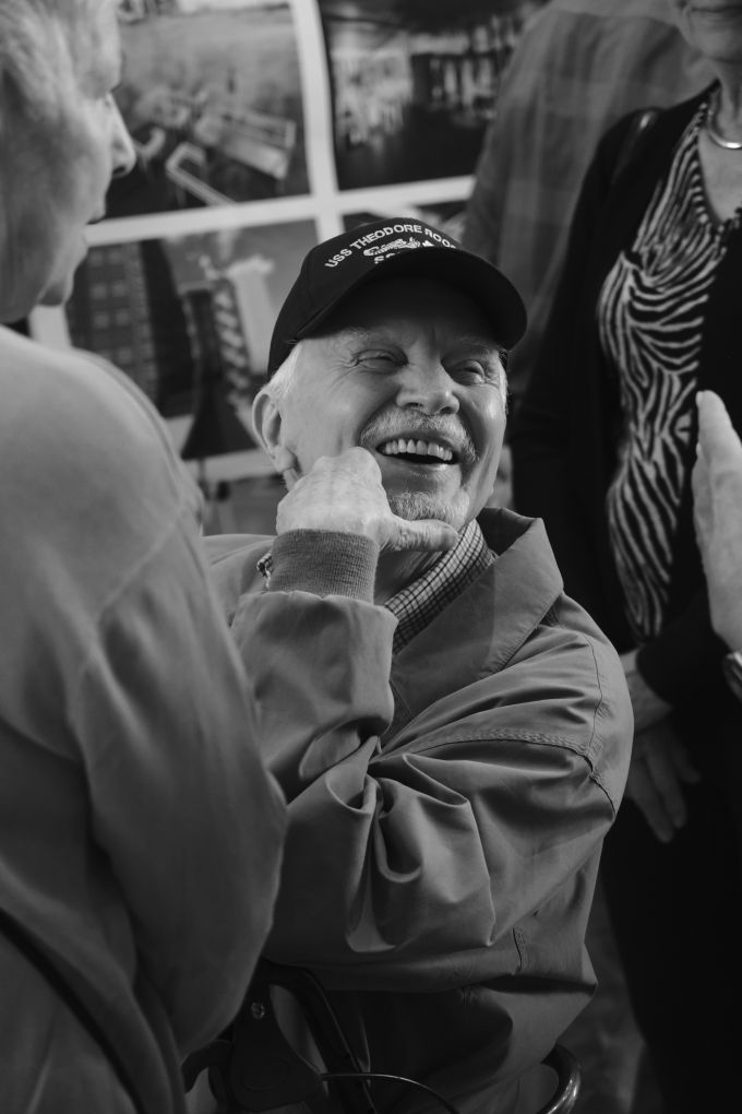 An elderly man wearing a USS Theodore Roosevelt cap, smiling among other attendees.