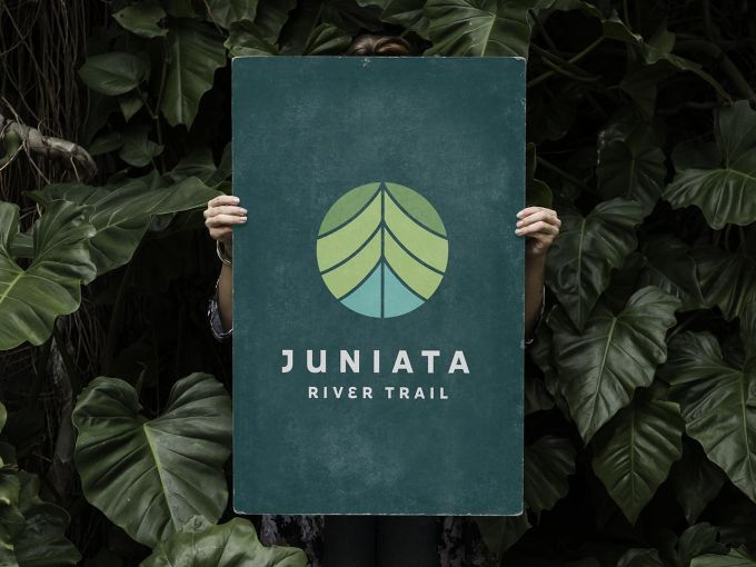 A person whose face is hidden behind a sign they are holding with a logo and the words Juniata River Trail on it.