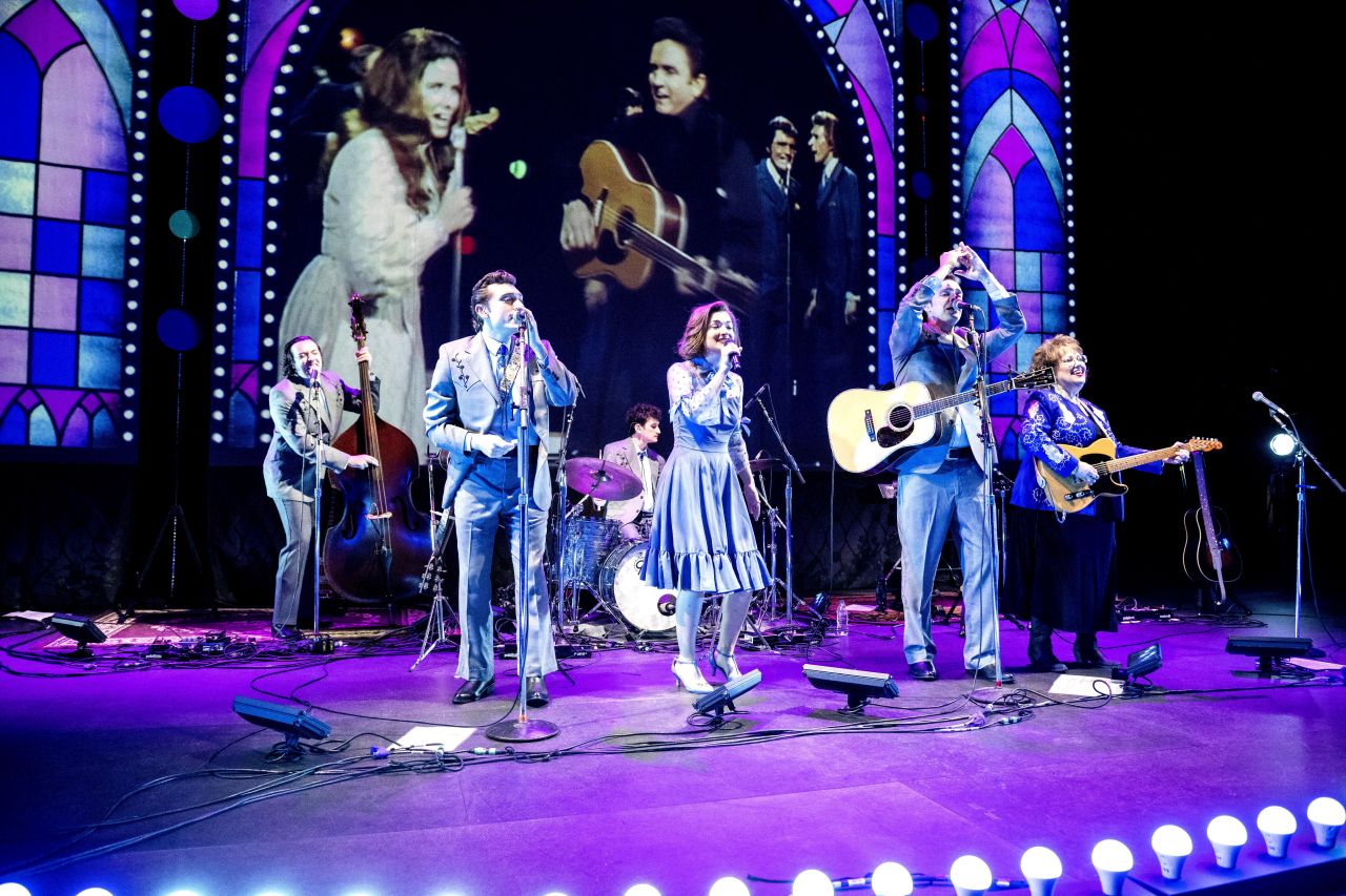 A band of six man and woman musicians perform on a stage while archival footage of Johnny Cash is screened behind them.