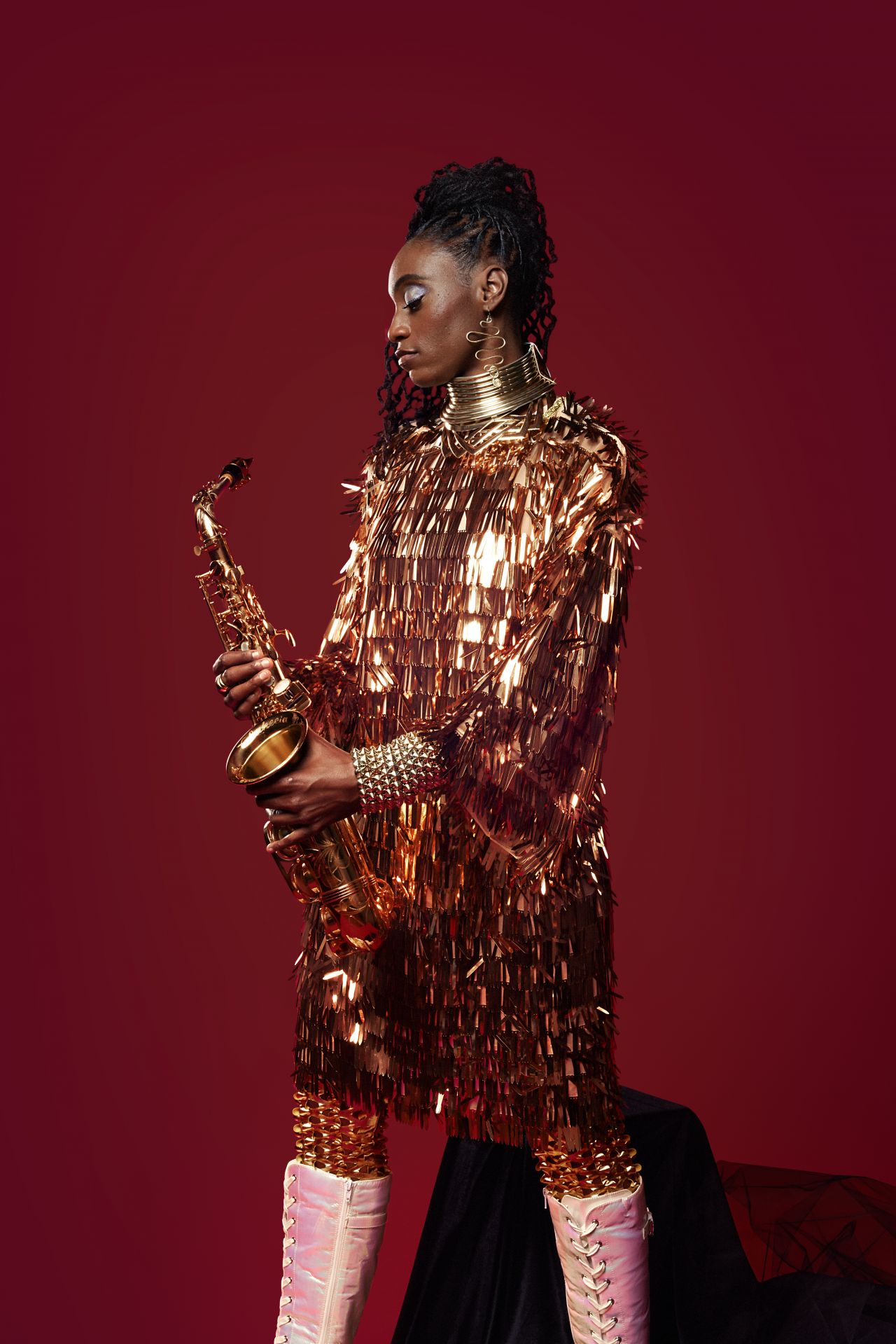 A young Black woman wearing her hair in dreadlocks and a shiny gold lamé cape holds her saxophone.