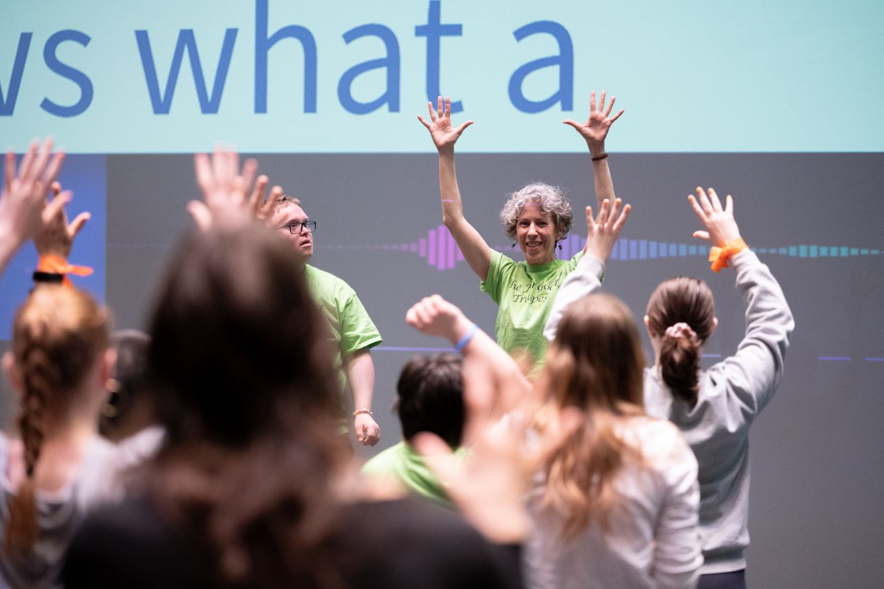 A woman instructing a group of people raises her arms high above her.