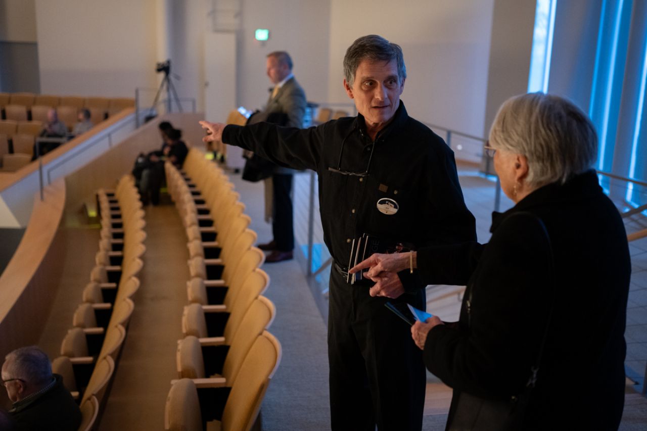An older man talking to a patron entering the auditorium points to a seating area.