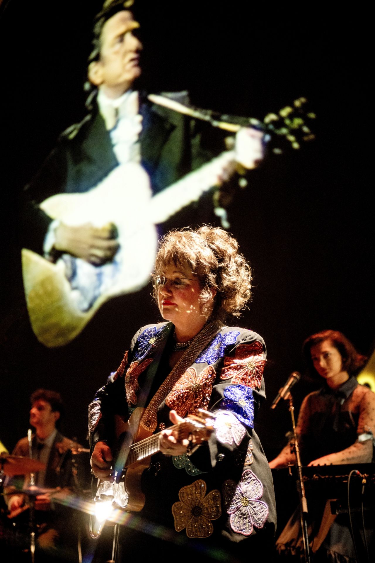 A woman wearing glasses with her hair pulled back plays a guitar on stage while archival footage of Johnny Cash shows in the background.
