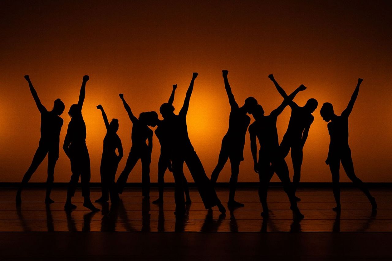 A line of dancers are shown in a silhouette against a dimly lit background collectively raising their fists.