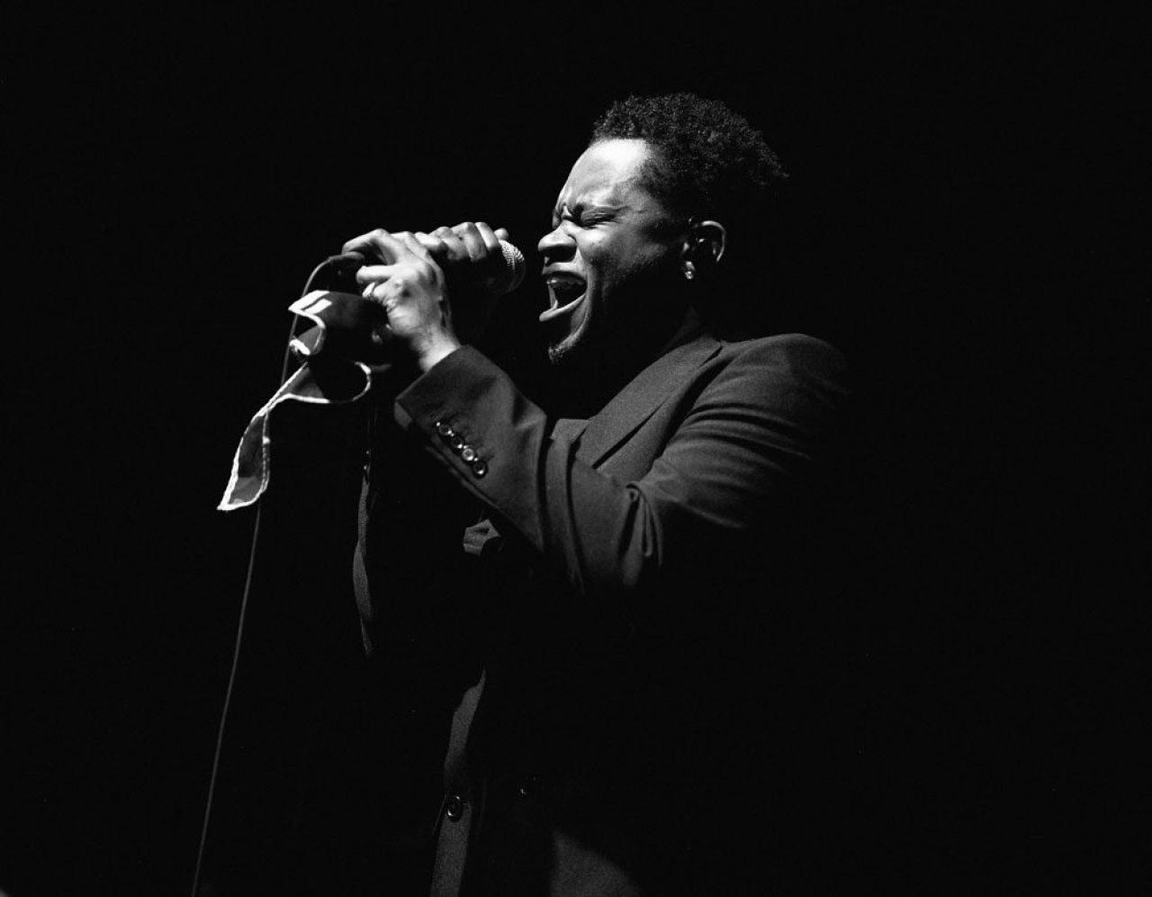 A Black man wearing a dark suit holds a microphone with both hands and sings into it.