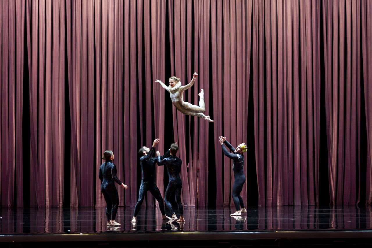 A group of acrobats wearing matching leotards throw and prepare to catch a woman dressed as a bird.