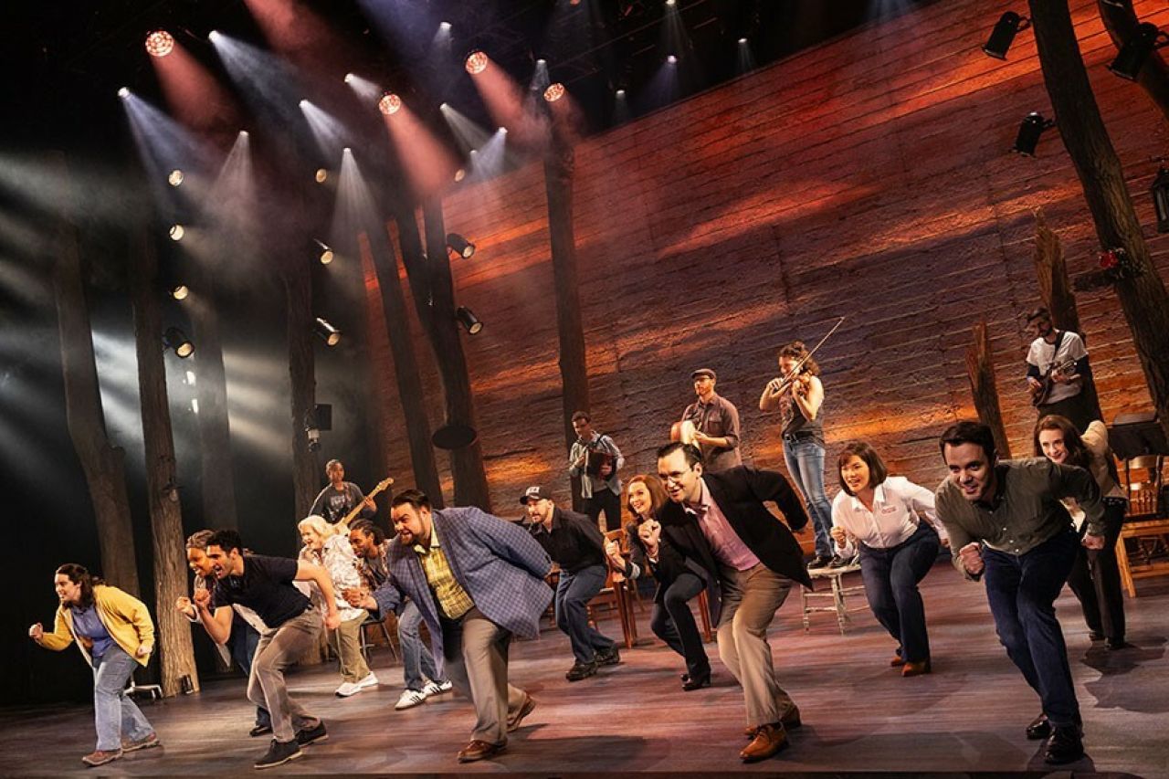 A diverse group of people of all ages sing and dance in unison on a stage.