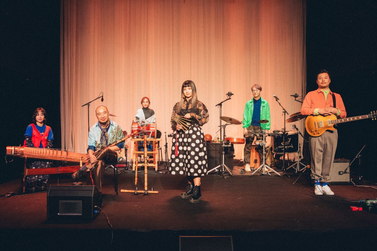 A group of Korean young adults stand on stage next to their rock instrument.