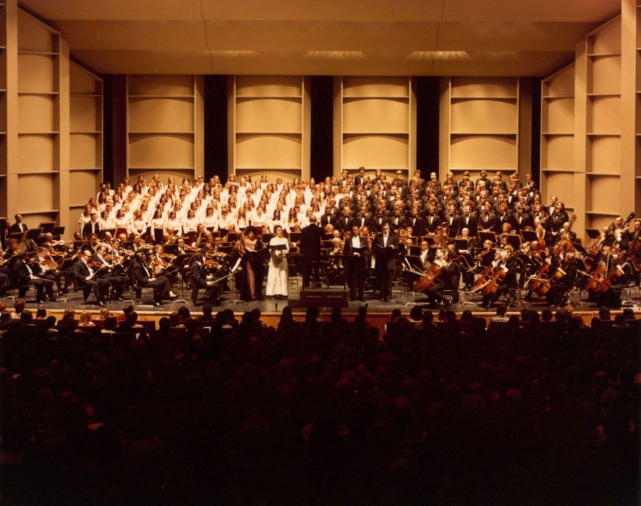 Numerous choruses and classical musicians stand in rows on a stage.