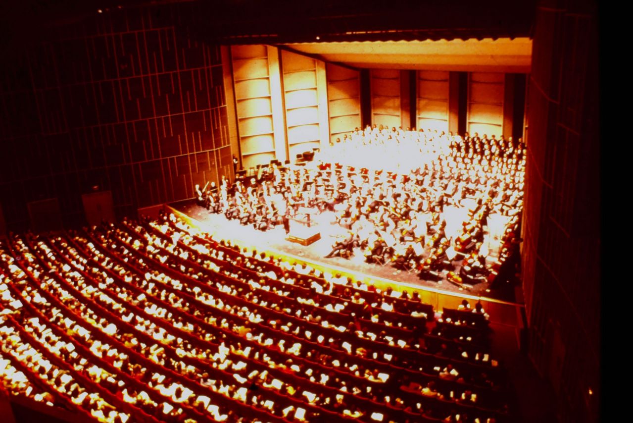An archival, bird'eye-view photo shows an orchestra audience watching musicians perform on the stage.