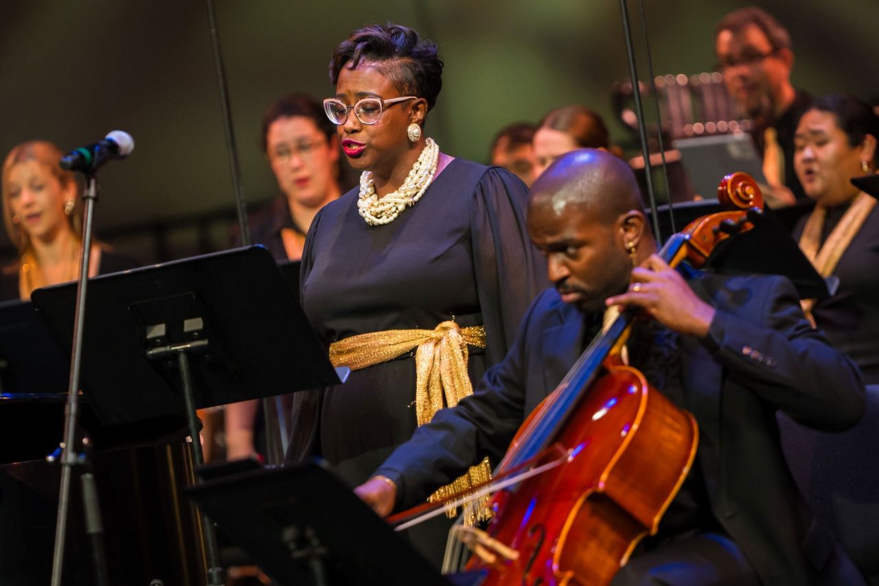 A diverse group of singers stand around a seated Black man playing cello.