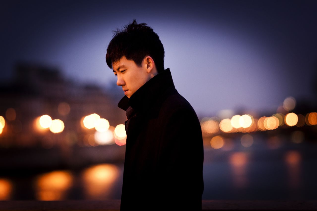 An Asian man wearing wearing a coat with the collar turned up while a cityscape at night is blurred in the background.