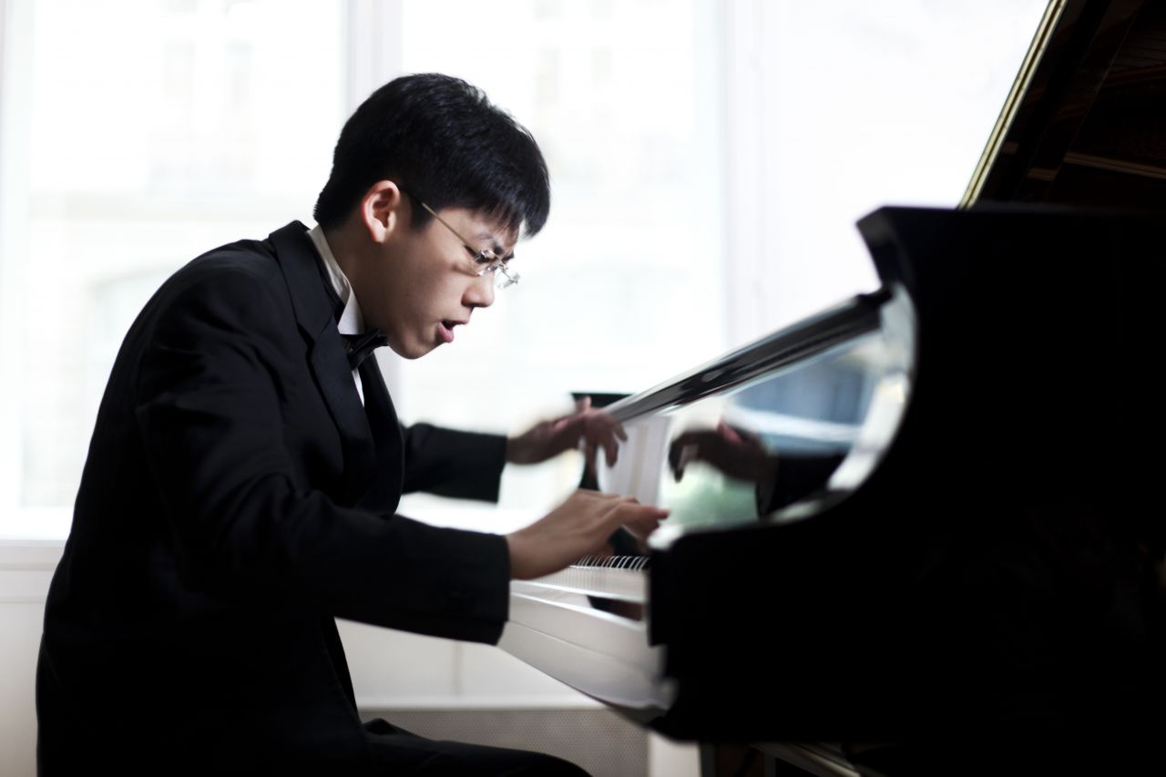 An Asian man wearing glasses sits at a piano and plays the keyboard with a look of intensity.