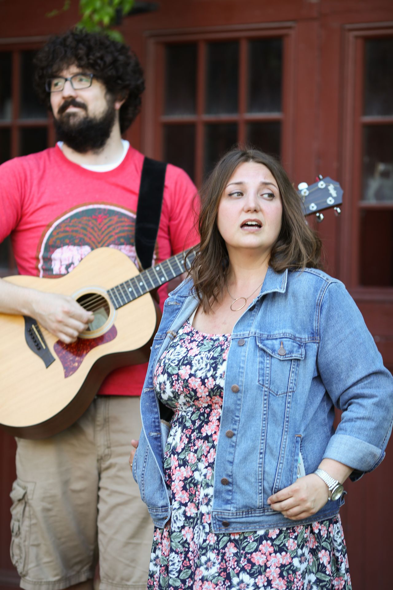 A man with a full head of curly hair and a bear strums a guitar while a woman standing next to him looks away.