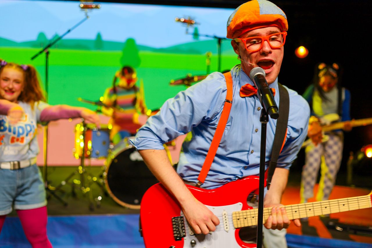 A boy wearing glasses plays a guitar in front of a diverse group of young people dancing behind him.