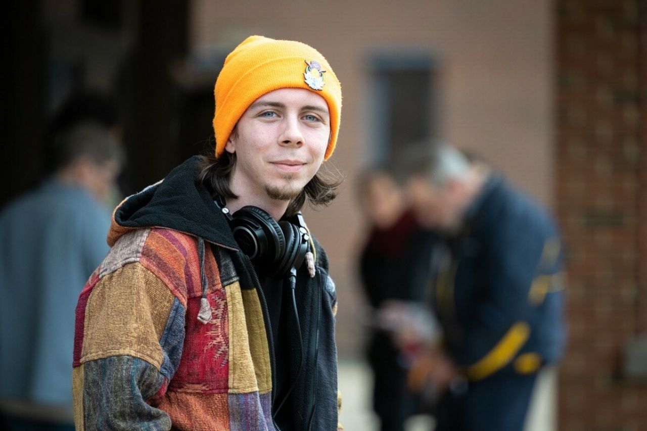 A young man wearing a knit cap, a patchwork jacquard jacket and large headphones around his neck smiles.