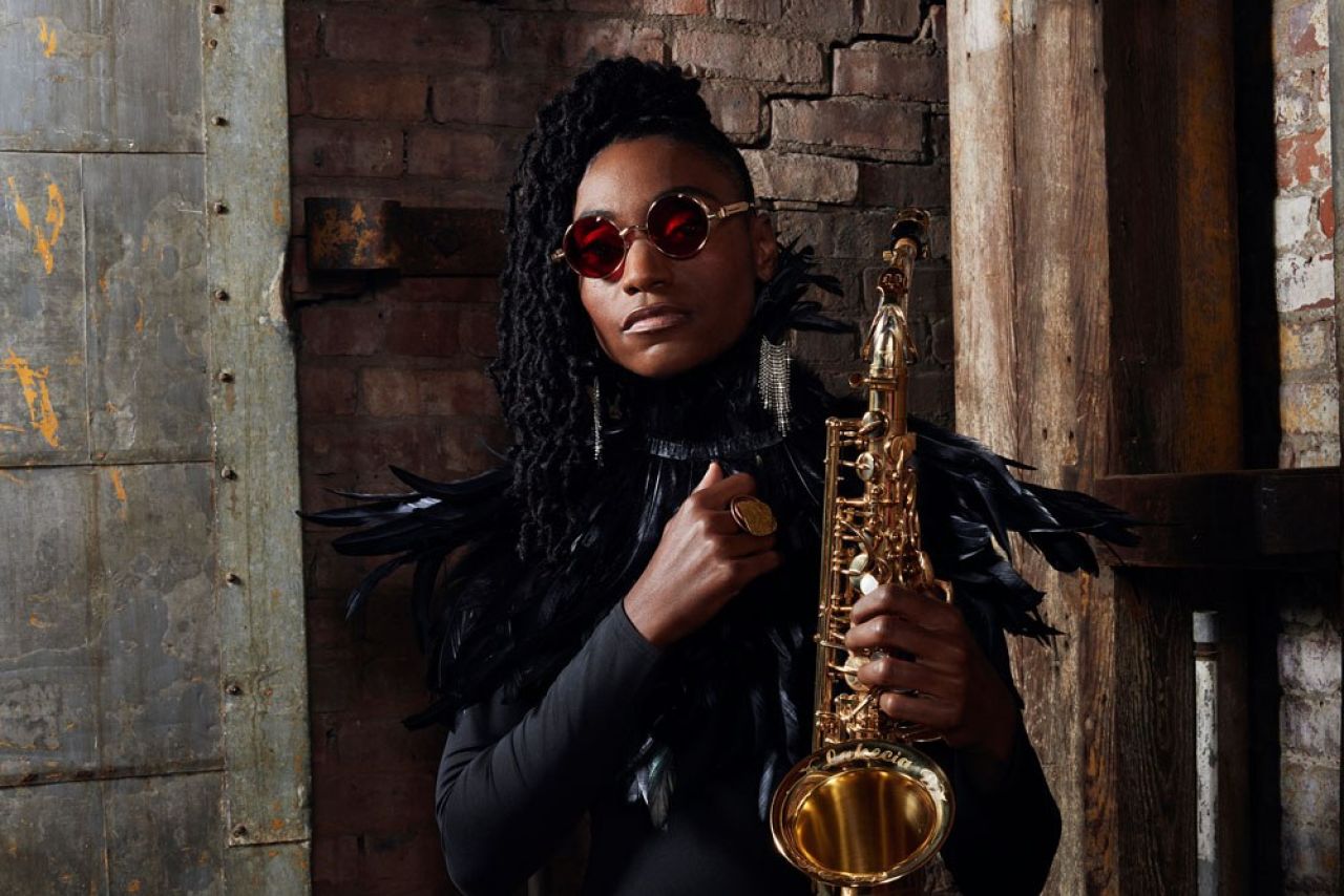 A Black woman wearing sunglasses stands against a brick wall and holds a shiny saxophone.