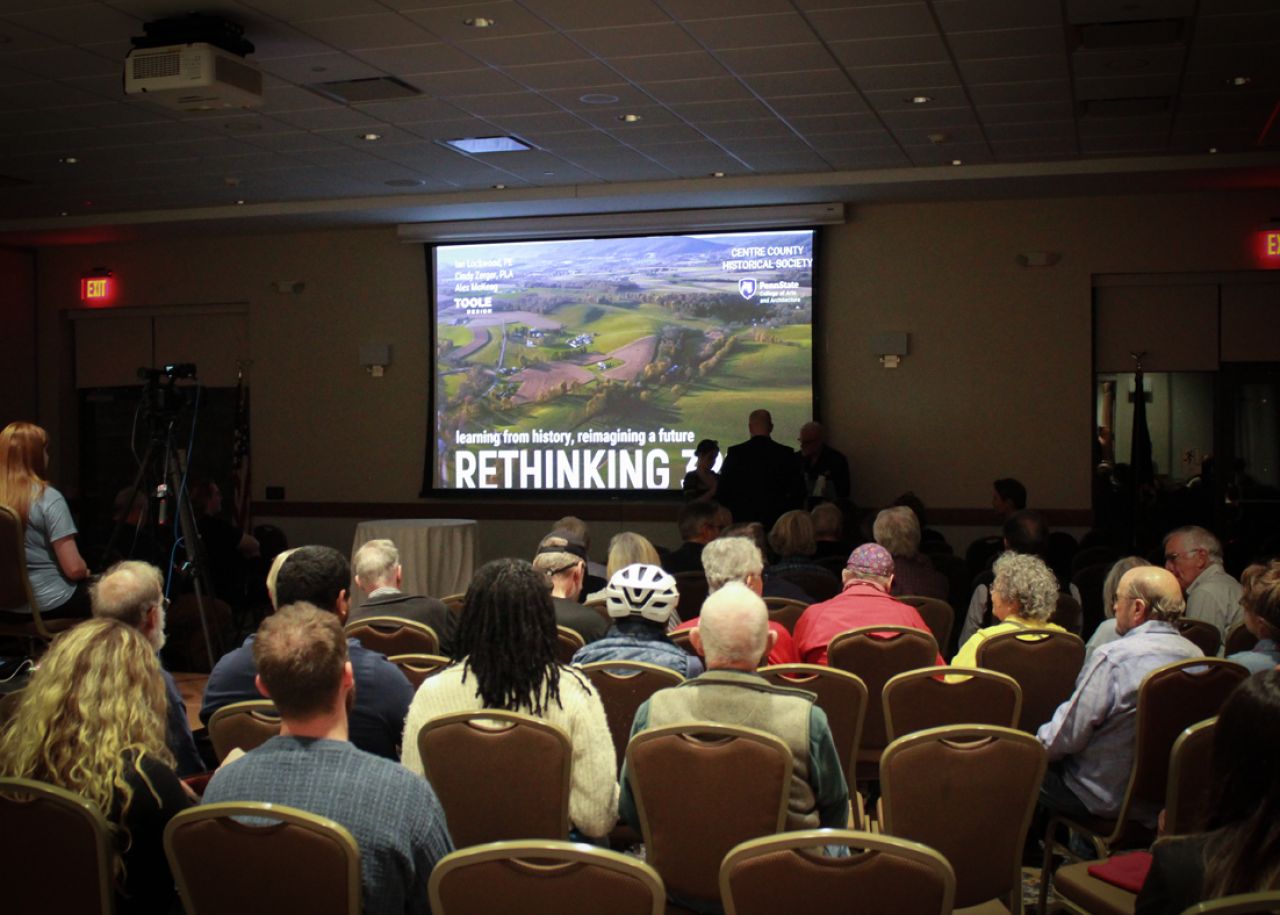 Looking into a conference room with people seated facing a screen featuring a slide that reads Rethinking 322.