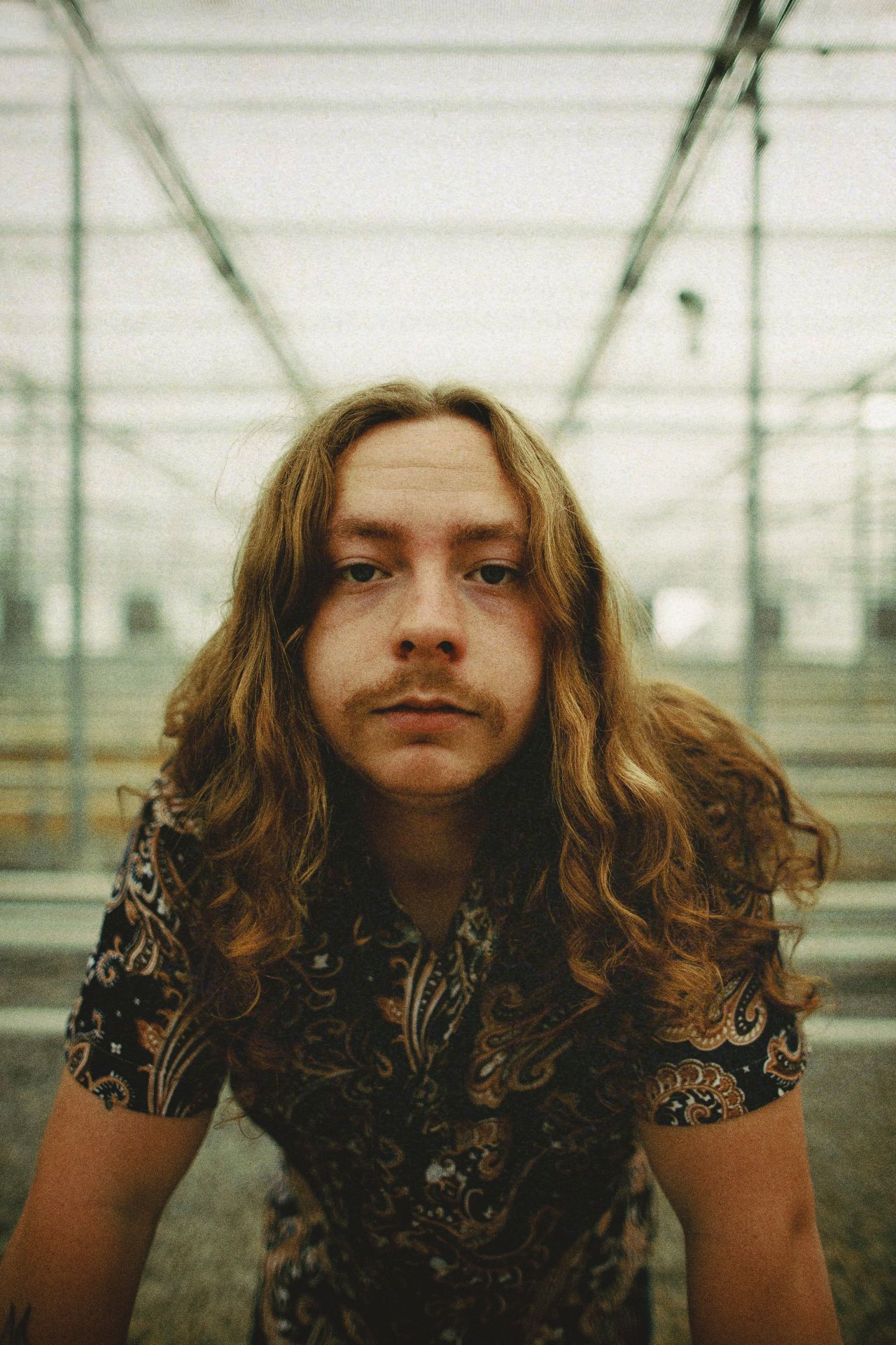 A young white man with long wavy hair leans against a table and looks at the camera.