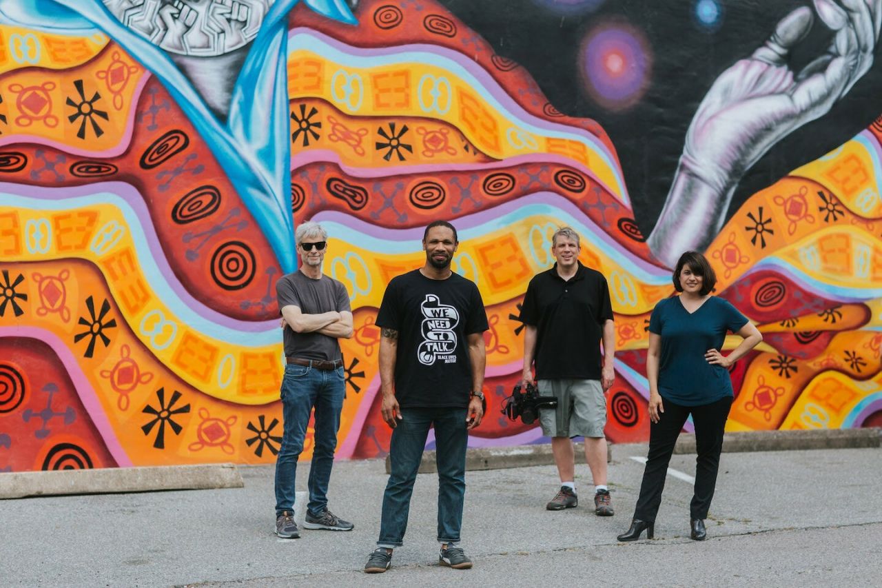 Four artists jauntily standing in front of a colorful mural.