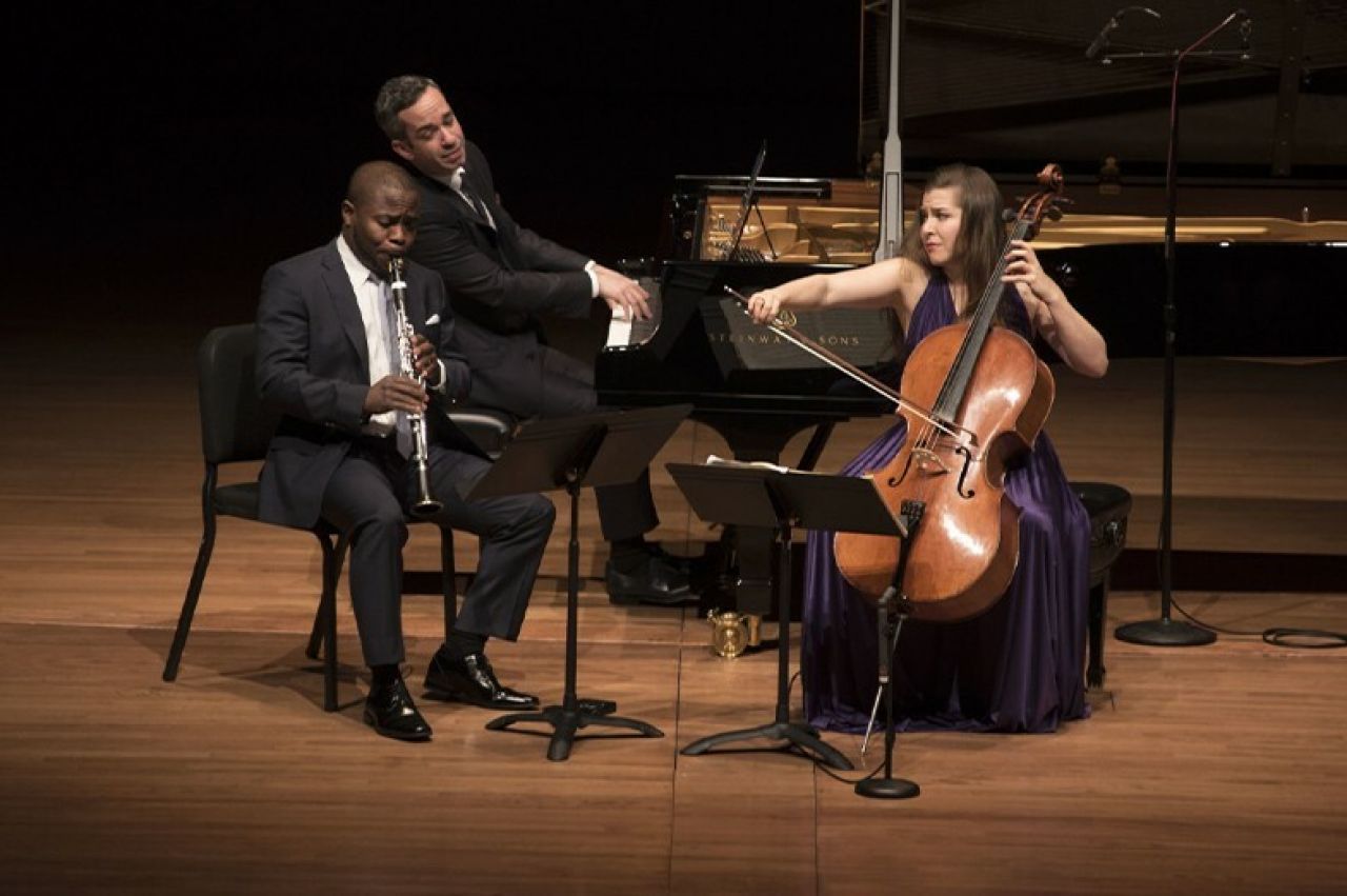 Three classical musicians sit in chairs on a stage and perform.