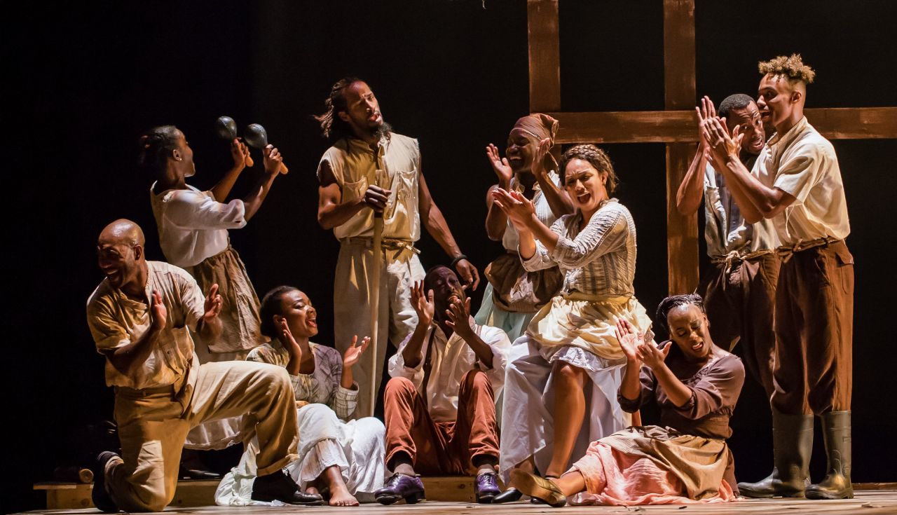 A group of about 10 Black artists wearing oversized and dirty clothing sit in a circle and clap their hands in unison.