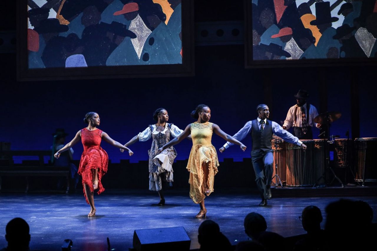 Black dancers dressed in 1930s dress dance in unison with their arms spread out and one leg lifted.