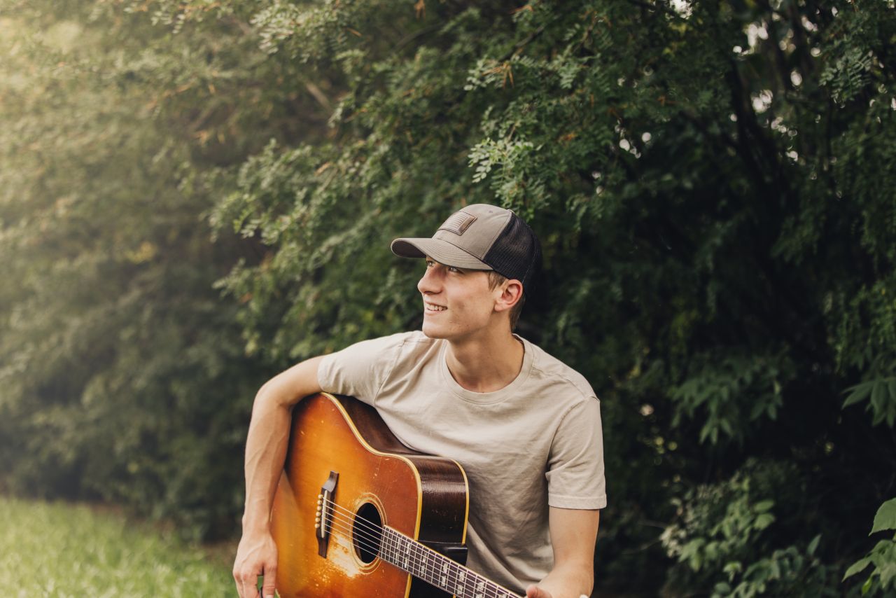 A young white man wearing a baseball hat rests an acoustic guitar on his lap and looks off into the distance.