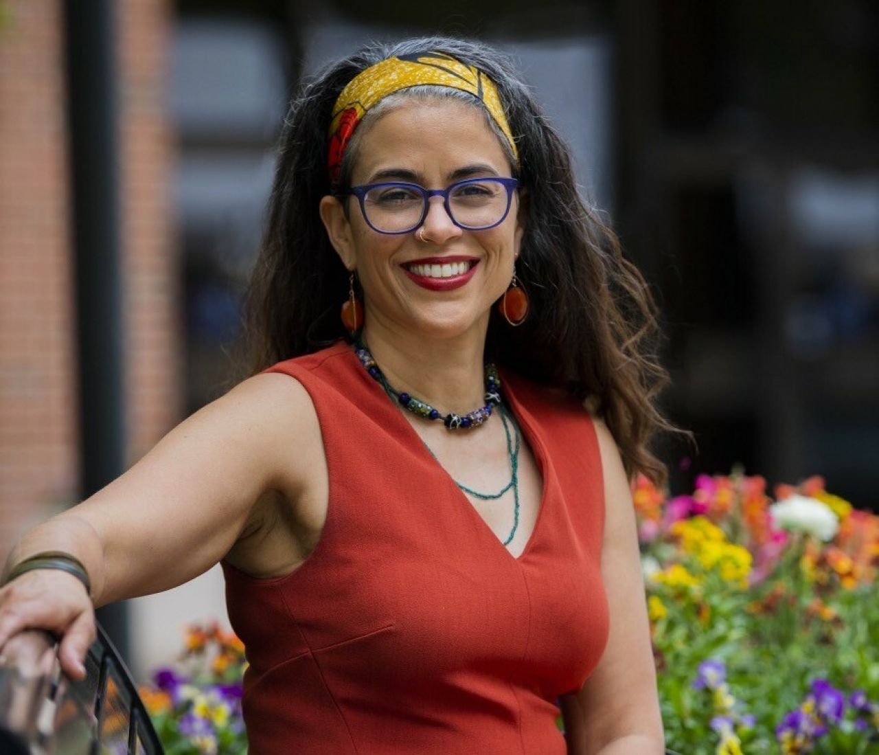 A woman smiles and rests her right arm on the back of a park bench.