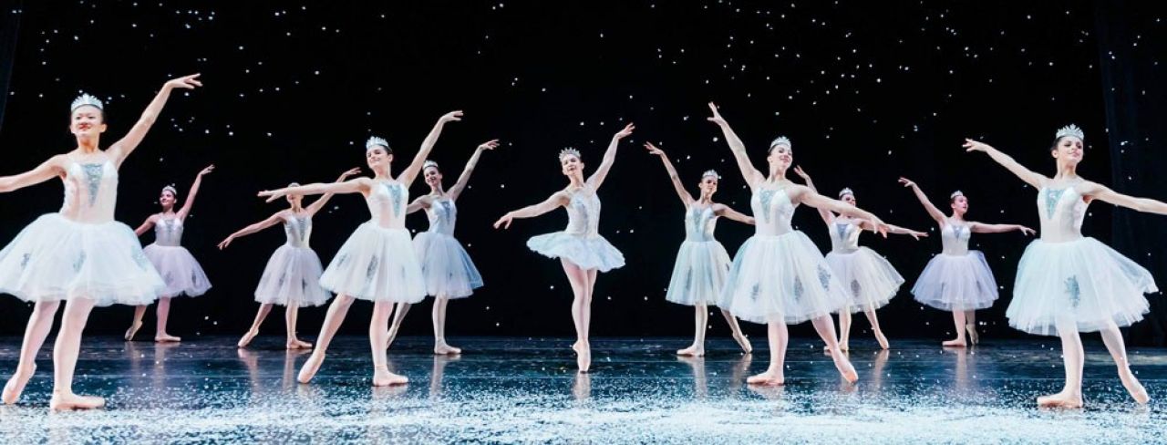 A group of young ballet dancers wear tutus and raise their arms above their heads.