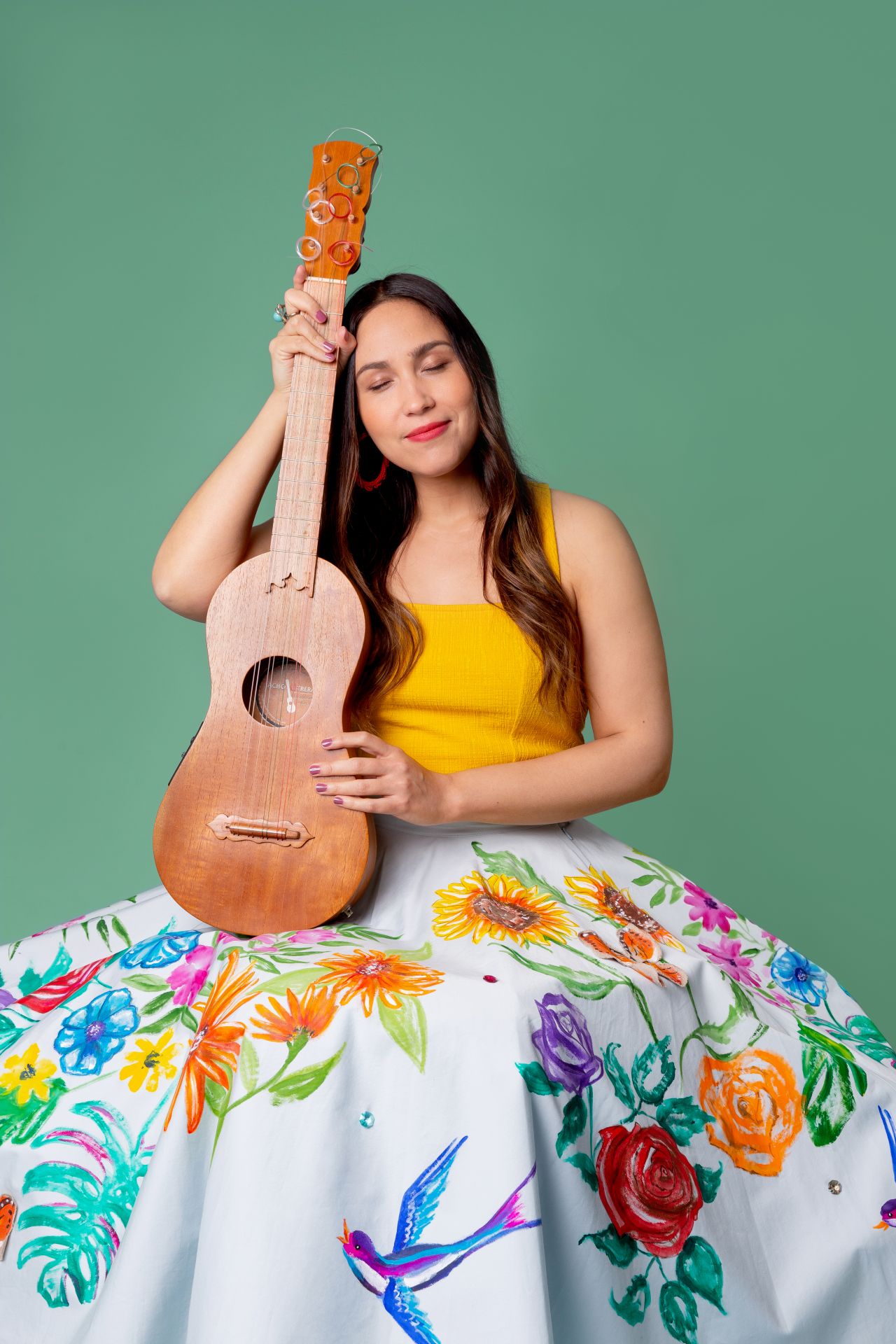 A smiling Latina woman wearing a full, embroidered skirt sits and holds a small guitar upright on her lap.