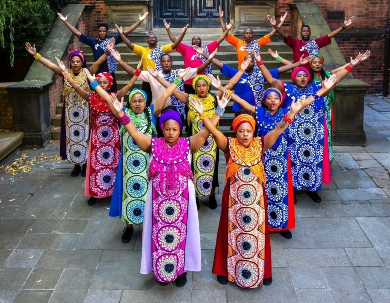 Artists wear boldly patterned robes and head scarves as they hold their arms up in a V formation.