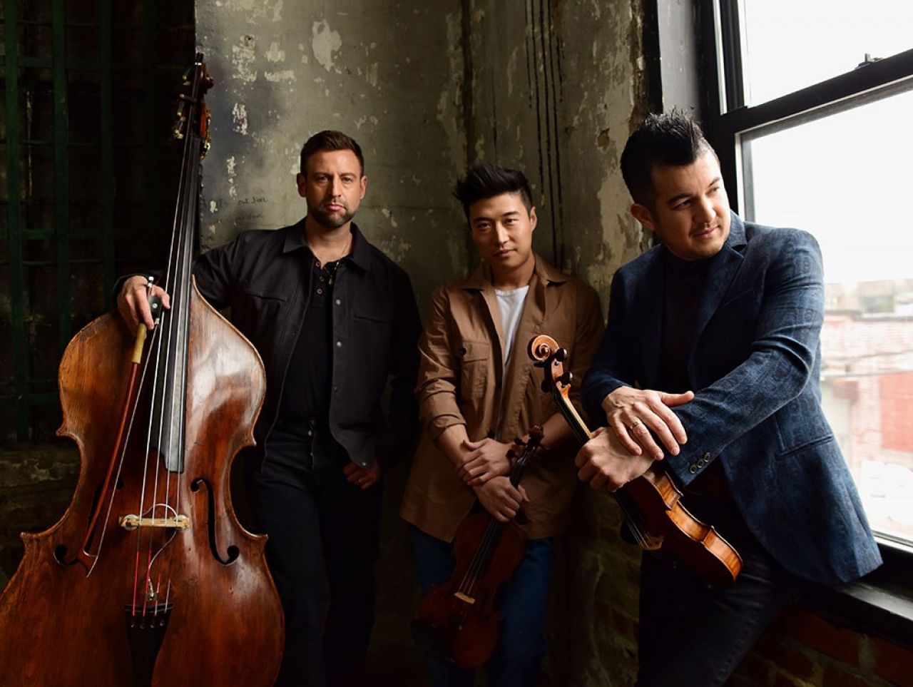 Three men stand next to a window in an urban landscape while they hold their string instruments.