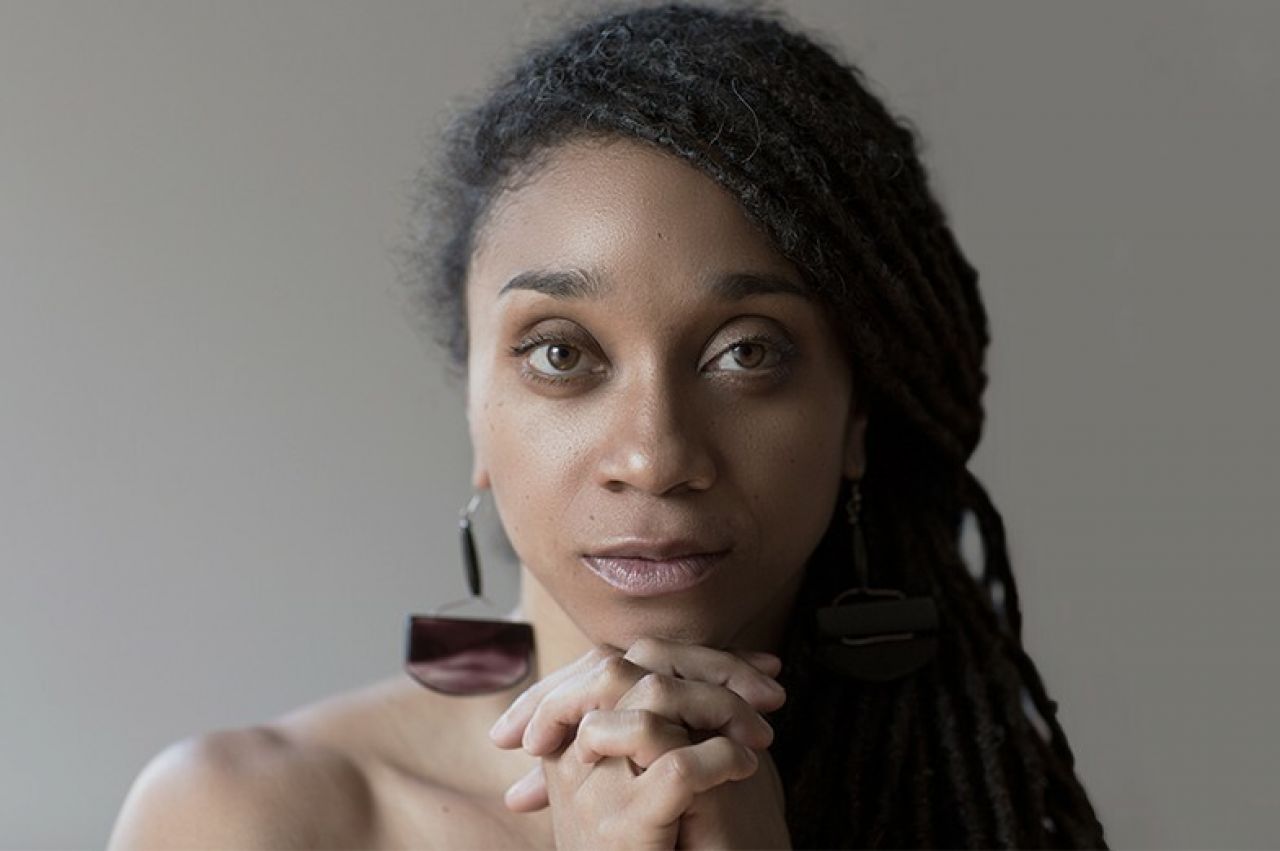 A woman clasps her hands at her chin and looks at the camera.